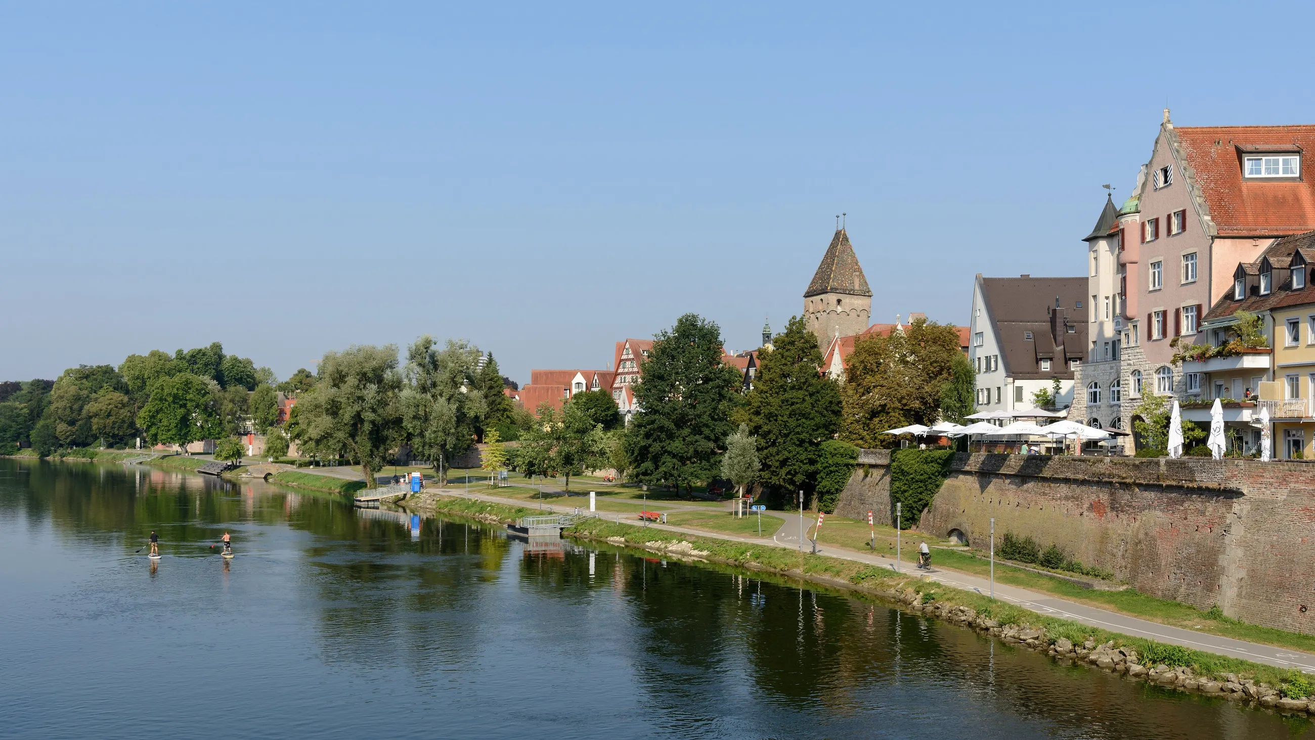 Die Donau in Ulm an einem klaren Tag, links das Ufer mit Bäumen, rechts die Stadtmauer und die Häuser der Altstadt
