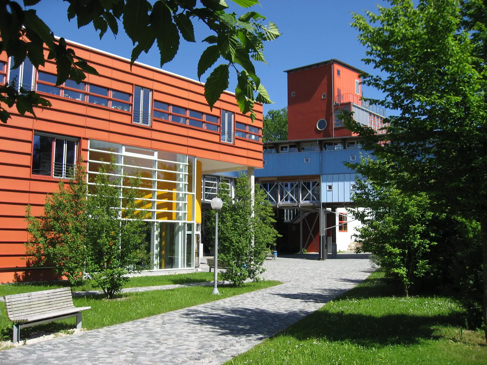 Die Bibliothek der Universität Ulm mit orangeroter Fassade und Fensterbändern, davor Bäume und ein Weg